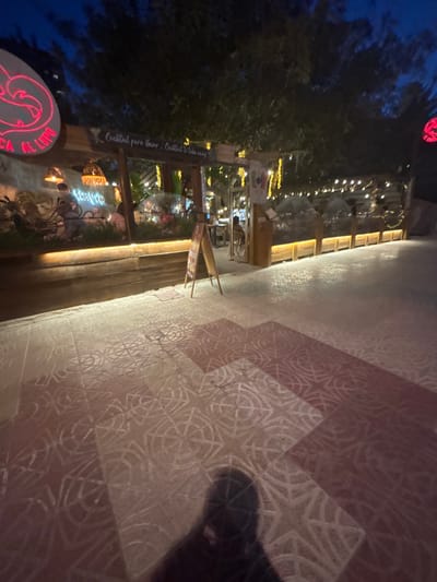 This media file captures a nighttime scene outside a building, likely a restaurant or bar, in Calp, Spain. The foreground is dominated by a textured tiled floor, with a repeating geometric pattern in shades of pink and white. A shadow of a person is cast on the floor to the right.
To the left, the scene opens up to a building facade with warm, inviting light emanating from within. String lights are draped across the exterior, creating a festive atmosphere. Trees with dark foliage are visible, some with their own decorative lights. A sign is mounted on a wooden post, with text that reads: "Cocktail para llevar / Cocktail to take away". Another illuminated sign, glowing in neon red and white, is partially visible at the bottom left, displaying the text "AL LUPO".
The time of day is clearly night, indicated by the darkness of the sky and the artificial lighting. The overall mood is lively and welcoming, suggesting a place of social gathering and enjoyment. The location cues like the Spanish text on the sign and the specific mention of Calp, Spain, further ground the image in a particular place. No specific event is discernable, but the setting suggests leisure and perhaps dining or drinking.