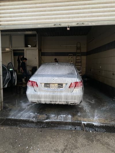 An image taken in Ma'raba, Syria, depicts the interior of a car wash bay with a silver Mercedes-Benz E 300 sedan centered, facing the viewer, and covered in white soap suds. The vehicle displays a Syrian license plate: "517 - SYR - 9920". Water and suds drain across a wet concrete floor into a metal grate system positioned in the foreground.

To the left, a male individual wearing a dark top and light pants stands near a dark-colored vehicle with its front passenger door open. On the right side of the Mercedes, partially obscured by the car's roof, a second male individual is visible. The interior walls of the bay are tiled with horizontal dark blue stripes. A tall, silver-colored metal ladder leans against the right wall. Modular white storage units are visible on the left wall behind the first individual. Overhead, a corrugated metal roll-up garage door is partially open, revealing a padlock hanging from its frame.