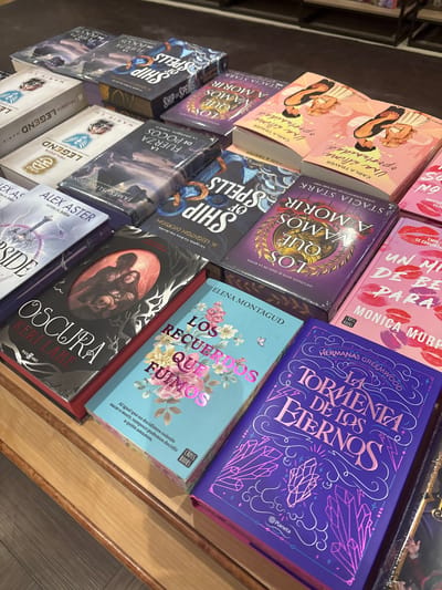 The image shows a selection of books displayed in what appears to be a bookstore or retail environment, likely in the city of Córdoba, Spain. The books are arranged on a wooden shelf and face upwards, revealing their cover art and titles in Spanish. Several titles are visible, including "Una Última Oportunidad" by Carla Traver, "A Morir Los Que Vamos" by Stacia Stark, "Ship of Spells" by H. Leighton Dickson, "La Fuerza de Pocos" by James Islington, "Legend" by Marie Lu, "Olvida" by Alex Aster, "Oscura" by Keri Lake, "Los Recuerdos Que Fuimos" by Elena Montagud, "Un Millon de Besos Para Él" by Monica Murphy, and "La Tormenta de Los Eternos" by Hermanas Greenmood. The books appear to be in good condition and are likely new. The lighting is bright, highlighting the details of the book covers. The wooden shelf is simple in design.