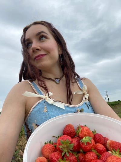 This media file features a young woman holding a white bowl filled with ripe strawberries. She is outdoors in what appears to be a field, with green foliage and a pole with wires visible in the background, suggesting a rural or agricultural setting. The sky is overcast with gray clouds.

The woman has long, dark brown hair and blue eyes. She is wearing a light blue dress adorned with embroidered birds. Her face is partially obscured by her hair and the angle of the photograph, but she has a gentle smile and appears to be wearing glitter on her cheeks. A necklace with a light blue stone is visible around her neck.

The activity taking place is the woman posing for a selfie while holding a bowl of freshly picked strawberries. The image evokes a sense of a pleasant summer day spent harvesting fruit. The lighting suggests it is daytime, possibly late afternoon or early evening, given the cloudy conditions. The location context is Кварса, Russia. There is no visible text in the image.