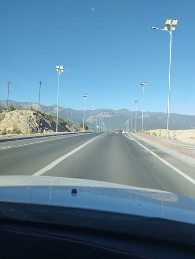 A wide-angle, eye-level shot captures a sunlit, paved highway receding into the distance, bordered by sparse vegetation and mountainous terrain under a clear blue sky. Solar-powered streetlights line the right side of the road, spaced at intervals. Power lines are visible on the left, supported by wooden poles. The foreground shows the hood of a vehicle, suggesting the image was taken from a car. The scene is devoid of people. The location of Myk, Uzbekistan, is not discernible from the visual elements present in the image.