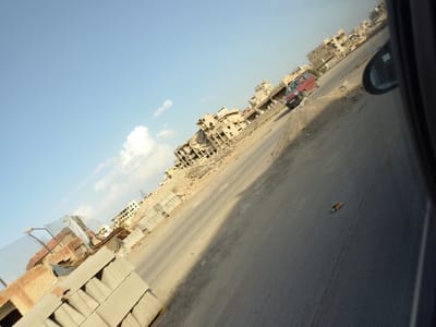 This diagonal wide-angle shot captures a scene of widespread destruction and desolation in Al-Qaboun Municipality, Syria, likely from the perspective of a vehicle in motion. The upper left portion of the image shows a clear, bright blue sky with sparse white clouds, indicating daytime conditions.

The central and right parts of the image depict a devastated urban landscape. Numerous multi-story buildings line what appears to be a dusty, partially paved road. These structures are severely damaged, reduced to concrete shells, piles of rubble, and twisted rebar, suggesting heavy shelling or demolition. In the mid-ground, a red, small car is visible on the road, partially obscured by debris and the uneven terrain, appearing to be parked or slowly moving past the ruins.

On the far right, the dark frame and reflective surface of a car's side mirror are visible, confirming that the photograph was taken from inside a vehicle. The road surface itself is rough, with visible cracks and scattered debris, including what looks like a piece of paper or packaging.

In the foreground on the left, stacks of concrete blocks and other construction materials or debris are piled up, hinting at either ongoing clearance efforts or the remains of previous structures. No people are visibly present, contributing to the desolate atmosphere. The overall impression is one of a post-conflict or disaster zone, with a sense of stagnation amidst the wreckage. No discernible text is visible in the image.