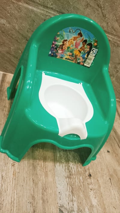 A green plastic potty chair with a white insert is positioned on a wooden floor. The potty chair has a sticker on the backrest featuring cartoon characters. The background consists of a tiled wall. The scene is indoors. There are no people visible in the image. The lighting appears to be artificial. There is no specific information to place this image in Onaldy, Turkmenistan.