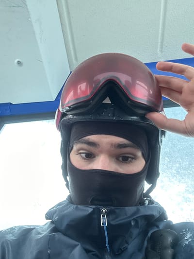The image depicts an individual, likely an adult male, captured in a close-up, low-angle shot. The person is wearing a black balaclava that covers the lower face and head, a dark ski helmet, and a dark blue waterproof jacket with a zippered front. Red-tinted ski goggles, exhibiting visible moisture droplets, are positioned on top of the helmet. A left hand, with a visible thumb and fingers, is seen holding the goggles. The individual's visible eyes are dark and focused slightly towards the viewer's right. The handle of a black ski pole is partially visible in the lower right corner. The background suggests an interior space, possibly a ski lift cabin, characterized by a white ceiling, a blue structural frame or railing, and a window on the left revealing a bright, undifferentiated outdoor environment, likely overcast or snowy. The image contains no specific visual identifiers to confirm the location as Anger, Austria.