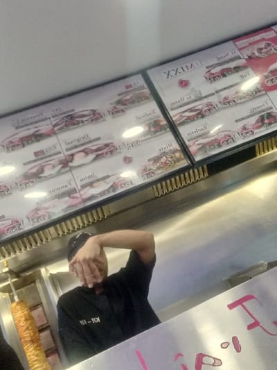 The image depicts the interior of a fast-food establishment, identified as a kebab restaurant, located in Sallaumines, France. A male worker, dressed in a black shirt with "KI-KI" visible on the front and a dark head covering, is positioned behind a stainless steel counter. His right arm is raised, with his hand obscuring his face. To the worker's left, a large vertical spit holds cooking döner kebab meat, which displays a golden-brown, cooked exterior. Above the counter, two illuminated menu boards are mounted, showcasing various food items, likely sandwiches or wraps, with associated prices and descriptions in French, including "XXL MIX," "Originel," "Raclette," and "Mixte." The foreground reveals a metallic counter surface with partial pink text or a logo, including the letter "F" and some dots. The scene is brightly lit, with reflections on the metallic surfaces and menu boards. The image is captured from a slightly low, tilted angle.