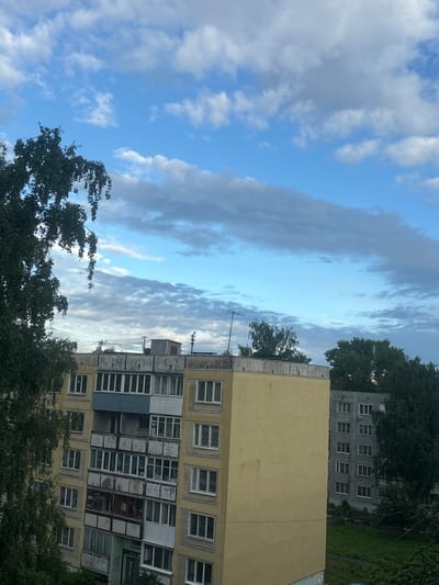 The image depicts a residential building in the city of Kirov, Russia, viewed from a slightly elevated angle. The building is predominantly yellow and has multiple floors with numerous windows and balconies. Some balconies appear enclosed with glass, while others are open. The architecture suggests a typical Soviet-era apartment block.

To the left of the main building, a large tree with dense foliage partially obstructs the view, its branches hanging downwards. Behind the yellow building, another apartment building, appearing to be gray, is visible, also with a grid of windows. Trees and greenery surround the buildings, indicating a residential area with some natural landscaping.

The sky above is a mix of blue and white clouds, with some areas of darker, possibly rain-laden clouds. Antennas are visible on the rooftops of some buildings. The overall impression is of a cloudy day in a residential district of a Russian city. There are no people or active events captured in the scene.