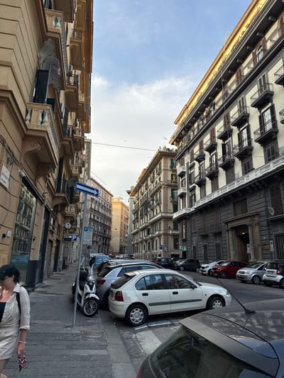 The image depicts a street scene in Naples, Italy. On the left, a woman wearing a straw hat, sunglasses, and a light-colored linen outfit walks along a sidewalk. She is carrying a patterned pouch in her right hand and has a bag slung over her left shoulder. The sidewalk is paved with cobblestones.
The street is lined with tall, multi-story buildings with balconies and shuttered windows. The architecture suggests an older European city. On the right side of the street, several cars are parked parallel to the curb, including a white sedan, a silver hatchback, and other vehicles. A white scooter is parked between two cars on the left side of the street.
Several street signs are visible, including a blue and white directional sign with an arrow pointing to the right, and a blue parking sign with the letter 'P'. The sky above is partly cloudy, with some areas of blue sky visible. The overall impression is a typical street scene in a European city during daylight hours.