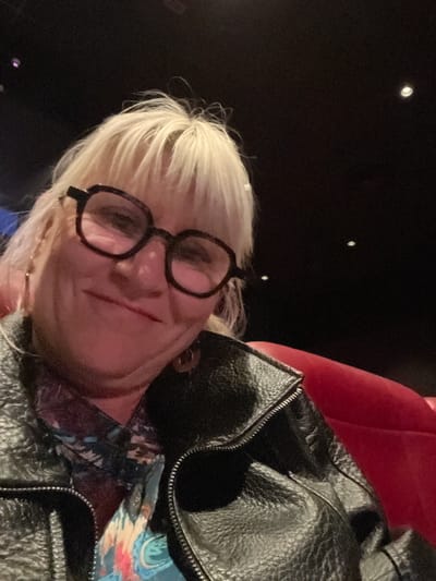 A person with short, blonde hair and wearing black-rimmed glasses is visible in the image. They are wearing a black, leather jacket. The background appears dark, with small, scattered points of light. A red object is partially visible in the background, likely part of a seating area. The image could have been taken indoors, possibly in a theater or auditorium. There is no definitive information to place the subject in Saint-Just-Saint-Rambert, France.