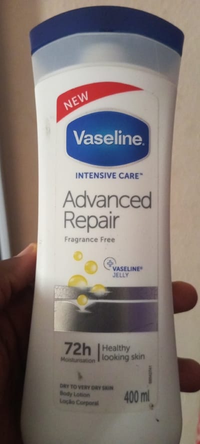The image shows a close-up of a Vaseline Intensive Care Advanced Repair body lotion bottle. The bottle is white with a blue cap and a blue oval logo containing the Vaseline brand name in white text. A red banner on the top left of the label reads "NEW". Below the Vaseline logo, "INTENSIVE CARE" is written in smaller text. "Advanced Repair" is prominently displayed in large gray text, followed by "Fragrance Free" in smaller text. A graphic of yellow droplets and a cracked surface is visible in the lower half of the label, along with a circular icon with a plus sign and the text "VASELINE® JELLY". Towards the bottom of the bottle, "72h Moisturisaton" and "Healthy looking skin" are printed. Further down, it states "DRY TO VERY DRY SKIN", "Body Lotion", and "Loção Corporal". The volume "400 ml" is visible on the right side of the bottle. A small serial number is also present on the right edge. The bottle is held by a person's hand, with fingers visible on the left and right sides of the image. The background is a neutral, light-colored wall. There is no information in the image that indicates the location is Baganakwo, Nigeria.