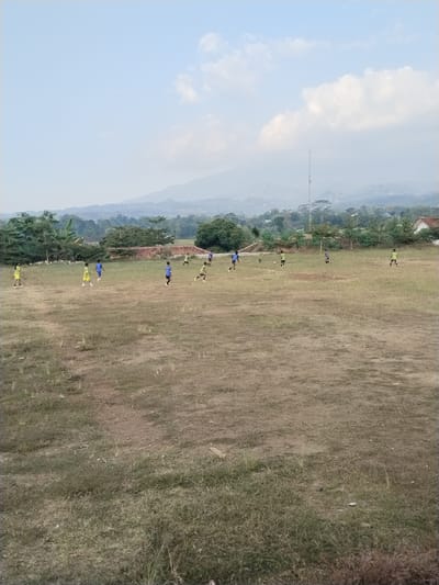 The image depicts an outdoor soccer game taking place on a dry, grassy field. Several young males, dressed in athletic attire including blue and yellow jerseys, are scattered across the field, some running and others appearing to be in possession of a soccer ball. The field is uneven and sparse in some areas, indicating a lack of regular maintenance. In the background, a line of trees and vegetation separates the field from a slightly hilly landscape. Further in the distance, houses are visible, suggesting a semi-rural or suburban setting. A tall antenna tower stands prominently against the horizon, accompanied by a hazy, mountainous backdrop under a pale blue sky with scattered clouds. The overall scene suggests a casual game played in a somewhat undeveloped area within the city of Kerjo, Indonesia.