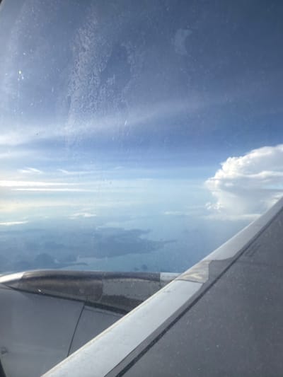 An aerial view from an airplane window shows clouds, sky, and land below. The wing of the airplane is visible in the foreground, as is the engine cowling. The land below appears to be a coastline with islands. The sky is a pale blue with some white clouds. The image is slightly hazy, likely due to dirt or smudges on the airplane window. There are no people visible in the image. The location is not identifiable in the image itself.