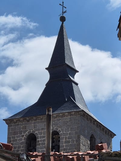 The image displays an upward-angled view of a church bell tower in El Espinar, Spain. The lower section of the tower is constructed from grey, rough-hewn stone blocks, featuring at least two arched bell openings. A dark bell is partially visible within the left archway. Above the stone structure, a dark grey slate spire rises, characterized by a flared, polygonal base transitioning into a taller, pointed pyramidal section. The spire culminates in a metallic finial comprised of a sphere, a Latin cross, and an arrow-shaped weathervane. In the immediate foreground, below the bell tower, are numerous reddish-brown terracotta roof tiles, some appearing stacked. A single, slender, light-colored cylindrical chimney pipe stands directly in front of the bell tower's lower portion, partially obscuring the left bell opening. The background consists of a bright blue sky with scattered white cumulus clouds. The scene is illuminated by direct sunlight, casting subtle shadows.