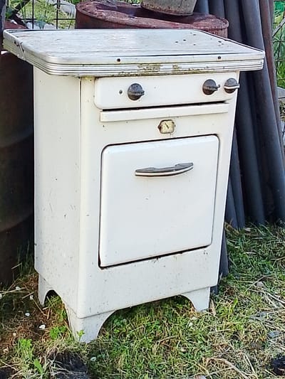 An old, white, two-burner gas stove with an oven is situated outdoors on grass. The stove has a metal top with chrome trim and two dark-colored control knobs. Below the burners is a drawer, and beneath that, a white oven door with a chrome handle. The stove stands on four curved, white legs. The background shows a metal fence, rusty barrels, and dark gray pipes. The environment appears to be a yard or outdoor storage area, with dry and green grass visible. The overall condition of the stove suggests it is no longer in use and has been exposed to the elements. The location is identified as Снопок Новый, Russia.