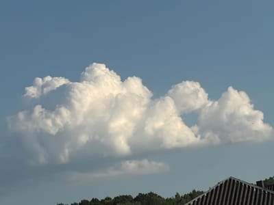 This media file captures a daytime scene over Hanniv, Ukraine. The primary subject is a large, fluffy cumulus cloud formation dominating the bright blue sky. The cloud is well-lit, suggesting it's midday or late afternoon, with sunlight illuminating its upper surfaces and creating shadows within its voluminous structure.
Below the clouds, a dark, wooded treeline stretches across the lower portion of the image. Peeking above the trees on the right side is the angular roof of a building, constructed with dark, ridged material, possibly asphalt shingles. The overall impression is one of a calm, natural landscape, with the sky and clouds being the focal point. The weather appears clear and pleasant, typical of a summer day. There is no visible text in the image.