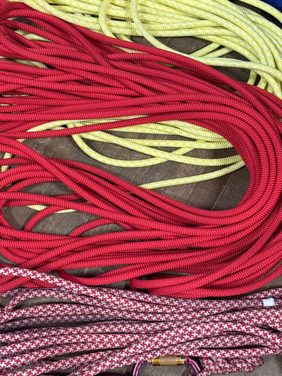 The image, reportedly captured in Tayshet, Russia, displays a collection of braided utility ropes laid out on a dark brown wooden surface, possibly a floor or table. The scene is devoid of people.
The top and central portions of the frame are dominated by a mass of loosely coiled, bright red braided rope. Interspersed with and beneath this red rope are sections of a yellow and white speckled braided rope. The lower section of the image features a distinct red and white patterned braided rope, characterized by a repeating 'plus' or houndstooth design. A metallic carabiner, featuring a purple body and a gold-colored screw gate, is partially visible at the bottom edge, attached to the red and white patterned rope. The wooden surface exhibits grain patterns and appears to be composed of planks. All objects are stationary.