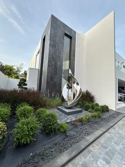 This media file shows a modern architectural exterior with a sculpted garden and a metallic sculpture. The building features clean lines with white and dark grey stone cladding. A prominent feature is a large, abstract metallic sculpture with a highly polished, reflective surface, positioned in the center of the garden. The garden itself is landscaped with dark mulch, dark grey pebbles, and a variety of green shrubs. Some ornamental grasses with reddish-brown plumes add texture and color. A narrow window is visible on the dark grey section of the building. In the background, a white structure with multiple levels and windows is partially visible, with a glimpse of a car parked in an open garage area. The scene appears to be outdoors during the daytime, with a clear sky. The location is identified as Baan Harn, Thailand. There is no visible text in the image. The overall impression is one of contemporary design and artistic expression.