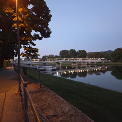 This image captures a tranquil evening scene along the Vah River in Piešťany, Slovakia. The foreground is dominated by a paved walkway with a metal railing, illuminated by an orange streetlamp and the warm glow from lights on a nearby bridge. Dark foliage from a large tree overhangs the left side of the walkway, its leaves catching the light. A few people are visible walking along the path in the distance.

The Vah River flows calmly beside the walkway, reflecting the lights of the modern, long bridge that spans it. This bridge features an enclosed section with numerous illuminated windows, suggesting it may be a pedestrian walkway or part of a building complex. Moored near the bank are two boats, one larger and white with "TRAJAN" visible on its side, and a smaller, more enclosed structure.

The background is a soft, twilight sky transitioning from a light blue to a deeper hue. Across the river, lush green grass slopes up towards a line of dense trees, with rolling hills or mountains visible in the far distance under the fading light. The overall atmosphere is peaceful and serene, with the artificial lights creating a warm contrast against the natural evening ambiance.