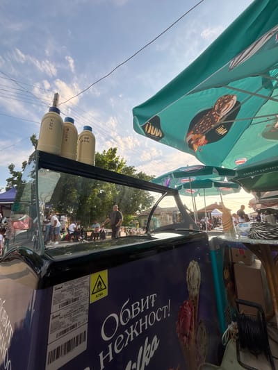 This detailed description outlines a vibrant outdoor market scene in Kostievo, Bulgaria, captured during daytime under a partly cloudy sky.
In the immediate foreground, a dark blue/purple ice cream display freezer is positioned at an angle. Its side features large white Cyrillic text, "ОБВИТ НЕЖНОСТ!" (Embrace Tenderness!) and "ilka", accompanied by a graphic of an ice cream cone containing vanilla and red scoops. A yellow triangular warning label with a flame symbol and "R290" is affixed to the freezer. A technical label lists "JUKA", "Model: Display Freezer H40SL", and other specifications.
On top of the freezer's glass display, three oversized, light-colored decorative bottles with blue caps are arranged. The leftmost bottle displays Cyrillic text "КЪПИ" (KAPI), the middle one "ЯГОДА" (YAGODA - Strawberry), and the rightmost "ВОР АМЕН" (VOR AMEN). The reflective glass surface of the freezer distorts the background elements.
Behind the freezer, a bustling area is visible. A man in a dark shirt and trousers stands near a small table, interacting with a group of casually dressed individuals, some seated and some standing. To the right of the freezer, a table holds a woven, pineapple-shaped decorative object and a black and white zebra-print bag.
Multiple large, teal-colored market umbrellas provide shade across the scene. The umbrella directly above the freezer is branded with a reversed "Nestlé" logo and an image of an ice cream bar, along with the Cyrillic text "ПЪРВАТА БЪЛГАРСКА" (THE FIRST BULGARIAN) and reversed "ICE CREAM YCHACH". Other similar "Nestlé" branded umbrellas are visible further back.
The upper portion of the image is dominated by a bright blue sky with scattered white cumulus clouds. Electrical wires crisscross the sky, supported by a utility pole. Green deciduous trees line the background, providing a natural backdrop to the market. In the distant background, low-rise buildings with tiled roofs, characteristic of the region, are faintly visible, and more people are dispersed throughout the area, indicating a lively community gathering or market event.