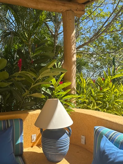 The image captures a vibrant, sunlit outdoor scene, likely a patio or garden, characterized by lush greenery and architectural elements. The perspective looks upwards and across, revealing a bright blue sky partially obscured by a dense canopy of trees and foliage.
On the left side of the frame, a sturdy wooden structure, possibly a pergola or arbor, is visible. It features a thick, rounded vertical wooden post connected to a horizontal wooden beam. The wood is a light, natural brown with visible grain, suggesting unfinished or lightly treated timber.
The central and left portions of the image are dominated by abundant green tropical or subtropical plants and trees. Various shades of green leaves create a rich texture, with some smaller, brighter red flowers, possibly from ginger plants, adding pops of color throughout the foliage. The sunlight filters through the leaves, creating highlights and shadows.
On the right side, a textured wall with a warm, sandy yellow or orange hue stands prominently. The wall has a rough, stucco-like finish. Mounted on this wall are two white electrical outlets, vertically aligned, with the upper one slightly higher. Below the upper outlet, a distinctive decorative element is affixed: a vertically ribbed, blue urn-shaped pot with handles, topped by a wide, inverted conical piece in a light, off-white color, resembling a lampshade or a large funnel. This piece is positioned with its wider opening facing outwards and downwards. Further down on the right, below the lower electrical outlet, part of a seating area is visible, featuring striped cushions in shades of blue and green, alongside a solid blue cushion, suggesting a comfortable outdoor bench or sofa.
The scene is brightly lit by strong sunlight, casting distinct shadows on the wooden beam, the foliage, and the textured wall, indicating a clear, sunny day, likely during the daytime. The overall impression is one of a tranquil and aesthetically pleasing outdoor living space.