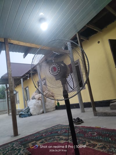 A floor fan with a silver grill is positioned in the foreground of an outdoor patio area. The fan is on a black stand and appears to be operating, as indicated by the blur of the blades. Behind the fan, a yellow building with a dark-colored roof is visible, along with wooden support beams and a white ceiling with lighting fixtures. Several large, white bags are stacked to the left of the building. A patterned rug covers the concrete ground in the foreground and extends towards the fan. In the background, more buildings and some greenery can be seen. The overall scene suggests a residential or communal outdoor space in Bokhtar, Tajikistan.