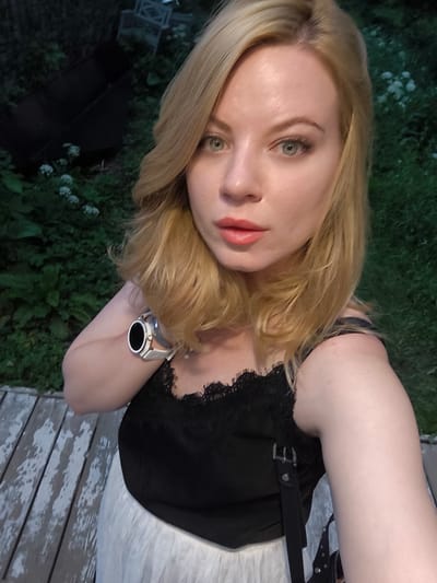 A woman with blonde hair and green eyes takes a selfie. She is wearing a black lace-trimmed tank top and a white skirt. A smartwatch with a white band is visible on her left wrist. She is standing on a wooden deck, with greenery and what appears to be outdoor furniture in the background. The setting is likely a residential area, possibly in Grebnevo, Russia, given the presence of lush vegetation and outdoor amenities. The lighting suggests it is daytime, likely late afternoon or early evening.