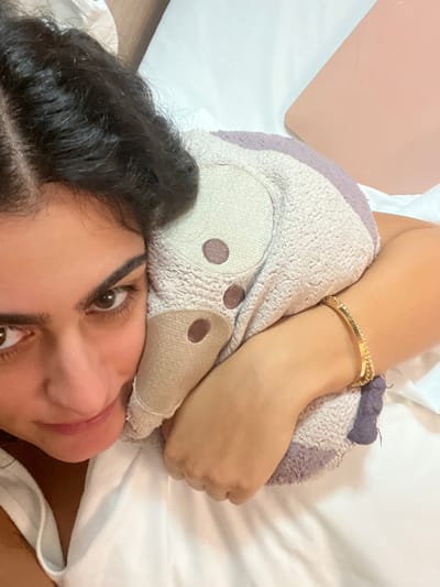 A close-up image depicts an individual lying on a white bedspread, positioned in an indoor setting within the city of Orléans, France. The subject is an adult female with dark, wavy hair, visible with a left part and subtle braiding near the scalp. She wears a white tank top or camisole and a gold-toned bracelet with a solid band and a thin chain on her right wrist. Her brown eyes are directed towards the camera with a neutral to slight smile expression. She embraces a plush, light-grey or beige stuffed object, which features abstract brown circular details for eyes and a snout, resting against her right cheek and chest. Her right arm is wrapped around the stuffed object. In the upper right background, a light pink or peach-colored rectangular object, possibly a laptop or tablet, is partially visible on the white bedding.
