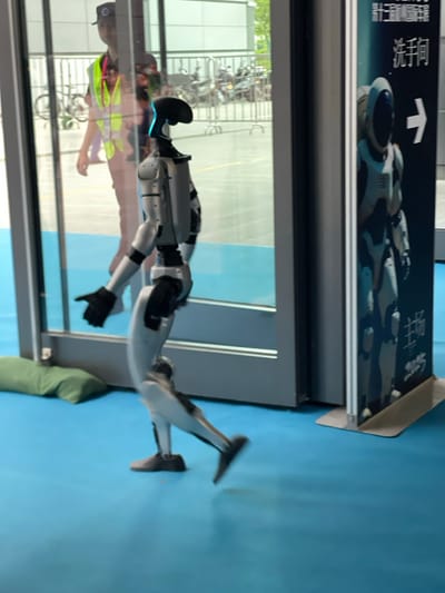A humanoid robot, painted white, walks out of a glass door, its left foot in motion.  A man, wearing a green vest and a blue cap, stands partially in the doorway and appears to be observing the robot. The scene is inside a building in Hangzhou, China. The location is near the West Lake Cultural Square and the coordinates are 30.272° N, 120.415° E. The floor is blue and a green cushion is on the floor.  The door leads to a hallway.  A sign, with Chinese characters, and an arrow pointing right, is to the right of the doorway.  The sign depicts a spaceman in a white spacesuit, standing in front of a dark blue background.  There are black metal railings in the background.