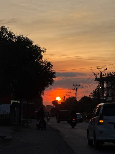 A street scene is captured at sunset in Palohlada, Indonesia. The sun, a vibrant orange sphere, is low on the horizon, casting a warm glow across the sky, which transitions from a pale yellow at the top to a deep orange and red near the sun. Silhouetted against the bright sky are power lines and utility poles, with one prominent pole with multiple crossarms positioned on the right side of the frame. A large tree with dense foliage dominates the left side, its dark form a stark contrast to the colorful sky. The road in the foreground is unpaved, appearing dusty or gravelly. Several vehicles are present: a white car is seen on the right, partially obscuring the view of the road; a dark SUV and a dark sedan are further down the road; and a few motorcycles are also visible. One motorcycle rider, wearing dark clothing, is positioned in the middle ground, moving away from the viewer. Another motorcycle with a passenger is further ahead. The overall atmosphere is that of a typical evening commute in a town or city.