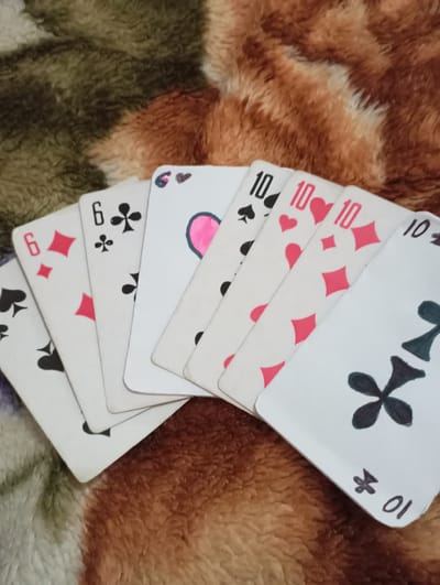 A hand of seven playing cards is fanned out across a plush, multi-toned surface displaying shades of brown, tan, and olive green. The visible cards, from left to right, consist of a partially visible black spade card, a 6 of Diamonds, a 6 of Clubs, a white card featuring a prominent pink heart symbol with a smaller black symbol above it, a 10 of Spades, a 10 of Hearts, a 10 of Diamonds, and a 10 of Clubs identified by a large black club symbol and the number '10' written vertically as '01'. The cards appear to be handmade or custom-designed, indicated by the drawing style of some symbols and numbers. No individuals are present in the image. This scene is situated in Kurganinsk, Russia.
