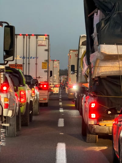A line of cars and trucks are stalled in a traffic jam. Red taillights glow in the dim light. Rain appears to have fallen as the road surface reflects the lights. The congestion features a variety of vehicles, from commercial trucks with white trailers to what appears to be smaller passenger vehicles. White lane dividers are visible on the road surface.