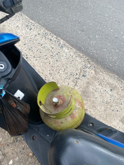 In Sungai Sipai, Indonesia, a gas cylinder sits on the floorboard of a scooter. The cylinder is primarily green with rust spots and a red top. A black plastic bag is tied to the scooter's handle with blue string. The scooter is parked next to a paved area with grey asphalt.