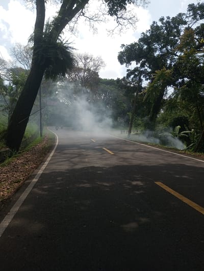A road in Nawng Hkar, Myanmar, is obscured by a large cloud of smoke. The road is asphalt with yellow lines marking lanes. Trees flank both sides of the road. Two motorcycles are visible in the distance through the smoke.