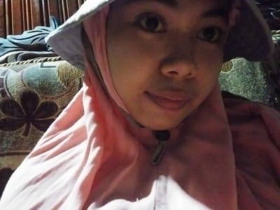 A close-up, eye-level shot shows a young woman wearing a light-colored bucket hat and a pink headscarf. She is looking directly at the camera with a slight smile. The background consists of a patterned sofa and a wooden structure. The lighting is dim, casting shadows on her face and the surrounding environment. The image appears to have been taken indoors. The location is identified as Labissa, Indonesia, though no specific landmarks are visible to confirm this.
