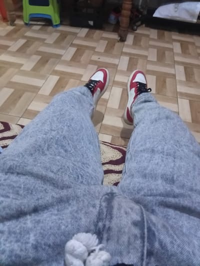 This image, taken in Martil, Morocco, presents a first-person, downward-facing perspective of a person's legs and feet indoors. The individual is wearing light-wash blue jeans or trousers, possibly with an acid-wash or faded texture, and stylish sneakers. The sneakers are predominantly white with prominent red accents on the sides and toe areas, complemented by black laces and some black detailing on the upper.
The person appears to be seated, with one leg resting on a patterned rug or cushion featuring dark red and beige/cream geometric patterns. The floor beneath is tiled with a distinct pattern of interlocking light and medium brown rectangular tiles, resembling a parquet design.
In the background, to the left, several indistinct objects are visible. These include a white bag or piece of fabric, on which the partial text "ro" can be seen. Below it, a dark object, possibly furniture, and a bright green and blue plastic item are visible on the floor. The overall scene suggests a casual, indoor setting, likely within a home. The lighting is ambient, offering no specific cues about the time of day.
