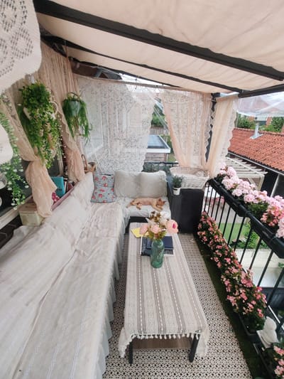 The image depicts a charming balcony in Hilversum, Netherlands, furnished for relaxation and adorned with plants and decorative elements. A long, pale couch with multiple cushions, one featuring a floral pattern, occupies the left side of the space. A tan dog is seen sleeping on the couch. In front of the couch is a rectangular coffee table covered with a lace tablecloth, holding a vase with pink and yellow flowers, a book, and a pen. The balcony is covered by a retractable awning, with sheer lace curtains draped along the sides, creating a soft, private atmosphere. Potted plants, including hanging greenery and pink flowers in planters along the railing, add vibrant color and life to the scene. The floor is covered with a patterned mat, and a black railing encloses the balcony, offering a view of neighboring rooftops and greenery. The overall impression is one of a cozy, well-decorated outdoor living space.