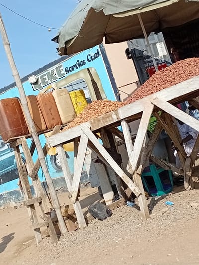 This image depicts an outdoor market or service area in Sani Mai Nagge, Nigeria. In the mid-ground, a crudely constructed wooden stand displays a substantial pile of light brown groundnuts. To the immediate left, four plastic jerricans, varying in color from reddish-brown to yellow, are arranged on an adjacent wooden support structure. The background features a light blue building wall painted with the text "KEKE service Cent." and a graphic depicting an auto-rickshaw. Above the scene, a weathered olive-green umbrella provides partial shade. The ground consists of dirt mixed with light-colored gravel, upon which a black plastic bottle and a teal plastic crate are visible. A tall, slender bamboo pole stands vertically on the far left. Electrical wires are present against the clear blue sky in the upper left. A person wearing white clothing is partially visible beneath the right side of the nut stand. Obscured signage near the right includes text related to "Money Trans" and "TRANS JEPA."