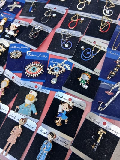 This image depicts a display of assorted brooches and pins, likely for sale, on a red surface. The items are presented in individual packaging, mostly black, with "Fashion Jewelry" printed on each. The brooches feature various designs, including medical-themed items like stethoscopes, doctors, and nurses, as well as an airplane, an evil eye, and decorative pins. The scene appears to be an outdoor market stall in Truskavets, Ukraine, suggested by the presence of these types of goods in such settings within the city. There are no people clearly visible in the image, and the focus is solely on the merchandise. The lighting suggests it's daytime, possibly under natural light.