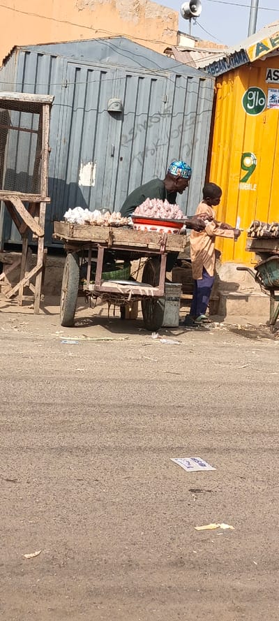 The scene depicts a street-level commercial area in Kaura Goje, Nigeria, under daylight conditions. Two individuals are present: an adult male and a child male.
The adult male, with dark skin and wearing a dark green long-sleeved shirt and a patterned blue and white cap, stands behind a two-wheeled wooden vendor cart. This cart displays multiple piles of light-colored, knobby produce, consistent with ginger or garlic, and a red-rimmed white enamel basin containing a reddish, granular substance.
The child male, with dark skin and dressed in a light tan tunic and dark blue trousers with green sandals, stands to the right of the main vendor cart. He holds a silver-bladed knife in his right hand, positioned towards dark, segmented stalks (likely sugarcane) arranged on a smaller, green metal cart.
In the background, two repurposed shipping containers serve as commercial structures. To the left, a weathered gray corrugated metal container exhibits partial white graffiti. To the right, a bright yellow corrugated metal container features prominent "glo" and "9 MOBILE" telecommunication logos, with the text "ABBAN UTAMA" visible above. Behind these structures, a faded orange-brown building wall with peeling plaster is visible. A white horn speaker is mounted on a pole above the yellow container.
The ground is a gray asphalt surface, sparsely scattered with small pieces of debris, including organic matter and a white paper wrapper with blue text in the foreground.