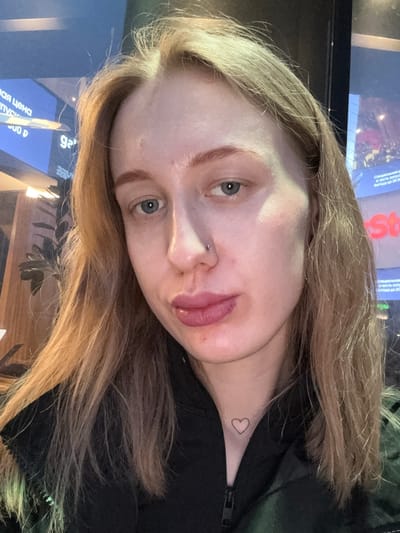 A close-up, rotated selfie of a fair-skinned young woman with light brown/blonde hair. She has a nose piercing and a heart tattoo on her neck. The background is slightly blurred and includes parts of screens displaying text in Cyrillic script. The background text includes “мая цена” and “Выгода до 20 00”. The image appears to have been taken indoors, possibly in a public space. There is no specific indication of Ol’ginskaya, Russia.