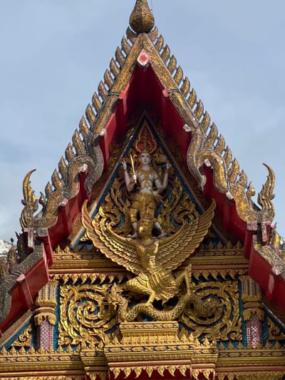 This is a close-up, eye-level shot of the ornate roof gable of a Buddhist temple in Chalong, Thailand. The primary focus is the detailed architectural decoration and a central statue. The gable is a triangular structure, characteristic of Thai temple architecture, covered in intricate gold and red embellishments. The apex of the gable features a statue of a deity with multiple arms, holding a sword-like object in its right hand. The deity is adorned in golden attire and stands atop a golden mythical bird with outstretched wings. Below the deity and bird, the gable is decorated with elaborate golden scrollwork, stylized dragon-like figures, and vibrant blue and red mosaic tiles. The sky above is a pale blue, with hints of clouds. The overall impression is one of grandeur and religious artistry.