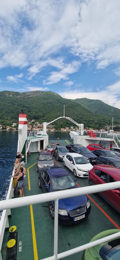 This media file depicts a ferry boat deck filled with cars, with a lush, green mountainside and a coastal town in the background. The scene is set against a backdrop of a bright blue sky with scattered white clouds, suggesting fair weather and daytime.
The primary subjects are the vehicles parked on the ferry's deck. There are several cars visible, including a red one, a dark blue Volkswagen with a Croatian license plate "HN BU963", a white car, and a light green car. Some of these cars appear to be occupied by passengers.
In the foreground, a group of people are visible, leaning over the railing of the ferry. They appear to be relaxed, enjoying the view or the journey. Their posture suggests a casual and leisurely atmosphere.
The setting is clearly a ferry operating on a body of water, likely a bay or strait, given the mountainous coastline. The presence of houses and buildings on the hillside indicates a populated area near the water. The location context of Lepetane, Montenegro, suggests this is likely the Strait of Verige.
The image is oriented vertically, with the sky at the top and the water at the bottom. The sun appears to be quite high in the sky, casting distinct shadows. There is some visible text on the ferry, including "ZABRANJENO PUŠENJE" which translates to "NO SMOKING" in Croatian, and "SWL 20T". The overall mood conveyed is one of travel and scenic beauty.