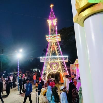 The image captures a lively night scene in Charfasson, Bangladesh, centered around a brightly illuminated, multi-tiered replica of the Eiffel Tower.

**Subjects:** The primary subjects are a large crowd of people of various ages, predominantly adults and youth, gathered around and walking past the decorative tower. Many individuals are dressed in traditional South Asian attire, with several women wearing hijabs or veils. On the far right, a large white and gold pillar, likely part of an entrance or structure, occupies the foreground. In the mid-ground, a street light pole wrapped with spiraling pink lights stands on the left. The dominant object is the illuminated tower, adorned with numerous colorful LED lights in pink, purple, yellow, and green. Decorative elements on the tower include a butterfly motif at its peak, a star in the middle section, and a large heart shape enclosing another star or triangle near its base. In the background, hints of other stalls or structures, also lit, are visible.

**Activity or event taking place:** The scene depicts a public gathering or festival at night. People are seen walking, observing the illuminated tower, and congregating, suggesting a festive atmosphere. The presence of such an elaborately decorated structure often indicates a local fair, celebration, or exhibition.

**Notable details:** The time of day is clearly night, with the dark blue sky contrasting sharply with the vibrant artificial lights. The clear sky suggests fair weather. The bright, colorful lighting of the tower and the street light creates a festive and energetic ambiance. The replica Eiffel Tower is a significant landmark in Charfasson, often lit up for special occasions. The crowd appears engaged and present, contributing to the bustling scene.

**Visible text:** On a sign in the mid-ground, near the base of the tower on the right side, there is visible text in Bengali. While partially obscured and difficult to read completely due to resolution, one word appears to be "একুরিয়াম" (Ekuriyam), which means "Aquarium," suggesting a possible attraction or stall at the event.