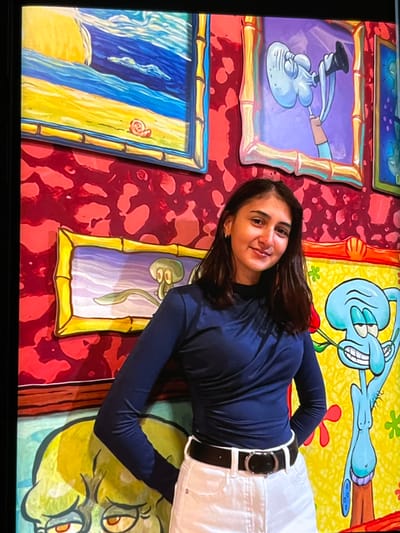 The image shows a young woman lying down on a colorful floor or wall decorated with cartoon images of characters from SpongeBob SquarePants. She is wearing a dark blue long-sleeved shirt and white pants with a black belt. Her body is positioned diagonally across the frame, with her head towards the upper right and her feet towards the lower left.
The background is a vibrant collage of cartoon art, featuring several framed pictures and abstract patterns. One prominent framed picture to the upper right depicts a smiling, blue octopus-like character, identified as Squidward Tentacles from SpongeBob SquarePants, in a dynamic pose against a yellow background with red flowers and green accents. Other framed images show variations of Squidward in different poses and settings, including one with a purple background where he appears to be holding a pipe-like object, and another depicting a beach scene with blue and yellow hues. A large, green cartoon face, likely another character from the show, is visible at the bottom right. The overall aesthetic suggests an indoor environment, possibly a themed attraction or exhibition space.
The woman is smiling and looking towards the camera, appearing relaxed and happy. Her hair is dark and falls around her shoulders. The lighting appears to be artificial, creating a bright and evenly lit scene. The colors are highly saturated and playful, consistent with the cartoon theme. There is no visible text in the image. The context of Nijmegen, Netherlands, may indicate this is a location within that city.