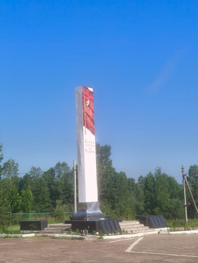 The image shows a tall, white obelisk monument with a red, shimmering panel adorned with a gold star on its upper portion. Cyrillic text is inscribed on the monument's lower section, reading "СЛАВА ПАВШИМ ЗА РОДИНУ" (Glory to those who fell for the Motherland). The monument is situated in an open, outdoor area with a dirt ground and a paved section marked with white lines. To the left of the monument, there are steps leading up to a slightly raised area with dark rectangular blocks. To the right, a wooden utility pole with wires is visible, and further to the right, more dark rectangular blocks are placed. A dense line of green trees forms the background, under a clear, bright blue sky. The scene is devoid of any people. The monument is identified as the monument to the fallen soldiers in the city of Kady, Kostroma Oblast, Russia.