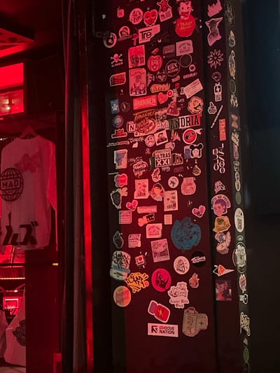 This image depicts an indoor scene dominated by a dark, vertical pillar extensively covered with numerous stickers. The environment is illuminated by a strong, uniform red ambient light, which casts a reddish hue over all visible objects.

On the left side of the frame, a merchandise display area is visible, featuring at least two white garments. The most prominent is a long-sleeved white shirt hanging on a dark hanger, adorned with a circular black graphic that includes the word "MAD" above a globe icon, with Japanese characters positioned below. A second white garment is partially visible beneath it. The red light originates from the left, highlighting the edge of a dark partition or wall that separates the merchandise display from the sticker-covered pillar.

The central and right portions of the image are filled by a dark, likely structural, pillar. This pillar's surface is almost entirely obscured by a dense, overlapping collection of diverse stickers of varying shapes, sizes, and designs. These stickers feature a mix of text, logos, abstract graphics, and stylized characters. Identifiable text on some stickers includes "Tre," "ABERMEN," "Andria," "Colectivo Ultras XXI," "Dianfres," "Support Your Local Dealers," "Happy Hippie," and "Groove Nation." The cumulative application of these stickers suggests a long-term accumulation of memorabilia or promotional materials.

Based on the explicit mention of "Colectivo Ultras XXI" (a known fan group of Millonarios FC, a football club based in Bogota) and the presence of what appear to be local event promoter or brand stickers like "Groove Nation," the location is precisely identified as being within Bogota, Capital District, Colombia.

The scene contains no individuals, focusing solely on the inanimate objects and the highly decorated environment, indicative of a subculture-oriented venue such as a bar, music club, or alternative retail space.