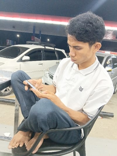 A young adult male is depicted seated barefoot in a black plastic chair, holding a silver smartphone with both hands, and looking down at the device. He is wearing a white short-sleeved polo shirt with a collar featuring black and white stripes, and a black chevron-like logo on the left chest. He has dark blue trousers on, with his right leg crossed over his left. The scene is outdoors at night, under an overhead canopy structure with prominent red and white banding, suggesting a gas station or covered parking area in Seumadam, Indonesia. In the background, a white Toyota Avanza equipped with black roof rails and a silver sedan are visible, parked on a paved surface under the artificial illumination of the overhead lights.