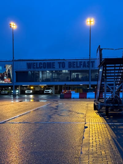 The image depicts the exterior of Belfast International Airport, located in Crumlin, United Kingdom, under twilight conditions. The main airport terminal building is visible, featuring large, illuminated white lettering that reads "WELCOME TO BELFAST" across its upper facade. Below this, the building has a level with multiple glass windows, revealing interior lighting, and a ground-level drive-through or covered parking area. Within this ground level, two white vans are parked, one with amber hazard lights active. A yellow service vehicle is partially visible on the far left. To the left of the building, a large illuminated advertising board displays "easyJet holidays" with an image of an archway.
The scene is lit by two tall metal light poles, each fitted with a cluster of four bright yellow lamps, casting strong illumination onto the foreground. The ground surface consists of wet asphalt with white painted lines and a section of wet, brick-patterned paving on the right. This wetness reflects the artificial lights. In the mid-ground, a series of red and white striped portable barriers are positioned. On the far right, a multi-tiered mobile aircraft boarding staircase or ramp is present, showing its structural components and wheels. No discernible individuals are present in the immediate foreground or mid-ground.