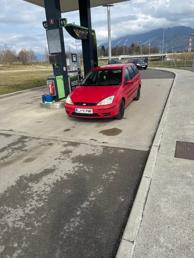 A red first-generation Ford Focus station wagon, bearing Slovenian license plate LJ 51-PMR, is positioned at a fuel pump beneath a dark grey and green canopy structure at a gas station in Radovljica, Slovenia. The vehicle faces slightly left of center. The fuel dispenser for lane 12, featuring a digital display and fuel nozzles, is directly adjacent to the car's driver's side. A red fire extinguisher in a stand and a blue plastic bucket containing a window cleaning squeegee are located near the pump. The ground beneath the vehicle and pump is concrete, displaying several dark, wet patches, and transitions to asphalt with additional damp areas. A concrete curb separates the main paved area from a raised walkway on the right. In the background, a large grassy field with bare deciduous trees extends to the left, while a range of snow-capped mountains is visible in the far distance under an overcast sky. A dark-colored vehicle with a small, indistinct figure nearby is seen further along an access road on the right side of the background. Utility poles and parts of a road overpass are also discernible in the mid-ground. No people are visible in the foreground.