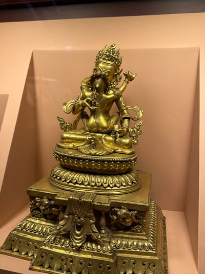 The image depicts a gilded statue, possibly of a deity or important figure, resting on an ornate base. The statue is richly detailed with intricate carvings. The surrounding environment appears to be a museum or gallery setting, judging by the neutral-colored wall and display niche. The statue is located in the city of Lalitpur, Nepal.
