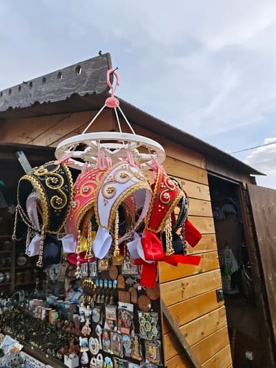 A market stall in Uglich, Russia displays handmade hats with ornate gold and silver beadwork. The hats are suspended from a white plastic hanger with pink clips attached to the side of a weathered wooden stall. Behind the hats, shelves are filled with souvenirs, ceramics, and other artisanal goods. The sky is visible above the stall, indicating an outdoor setting.